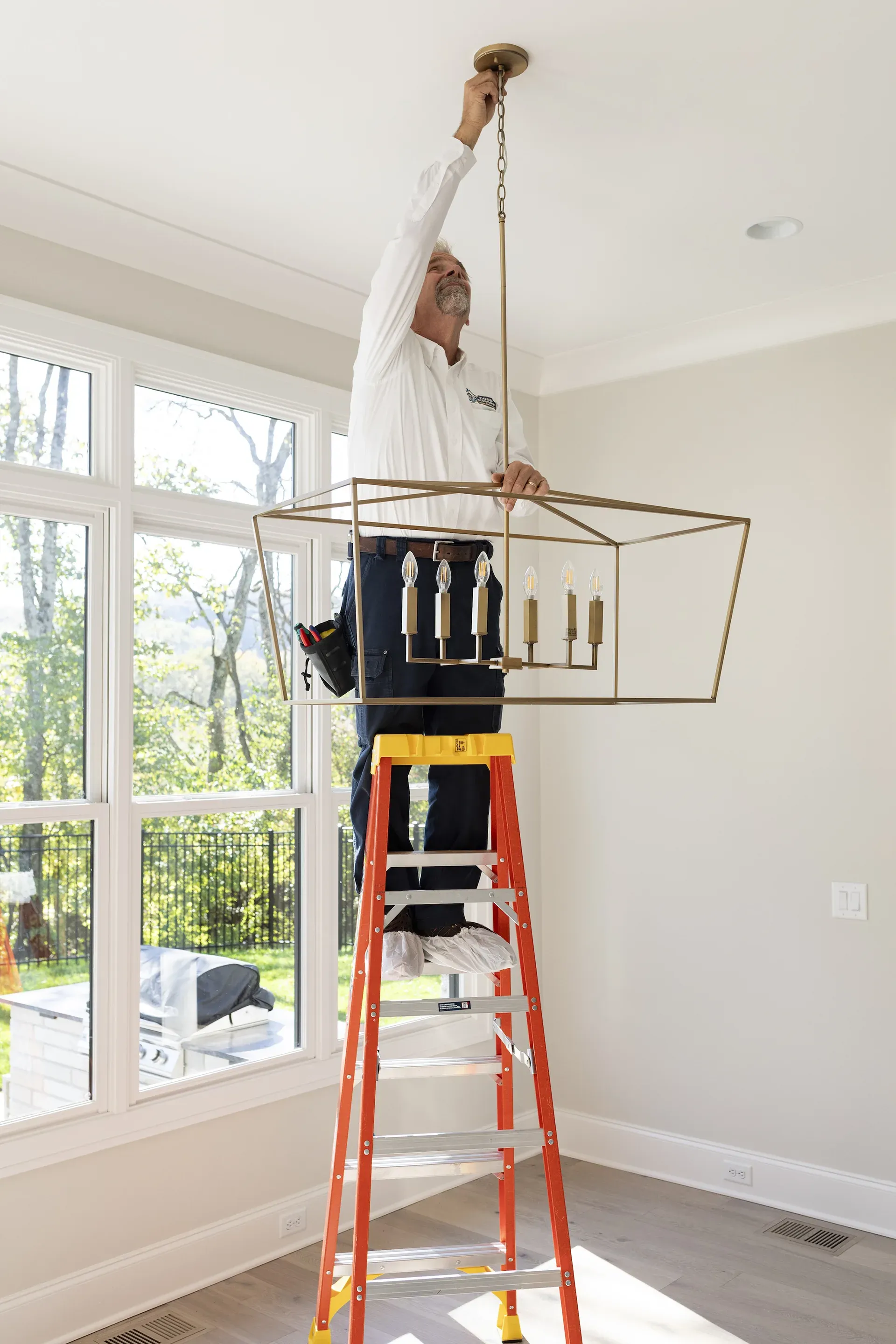 Man on ladder installing a light fixture in a room with large windows.